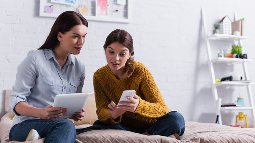 Mädchen hält Smartphone in der Nähe der Mutter mit digitalem Tablet auf dem Bett