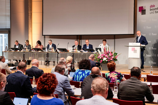 Eine Gruppe von Menschen sitzt an Schreibtischen. rechts im Bild steht ein Mann hinter einem Rednerpult