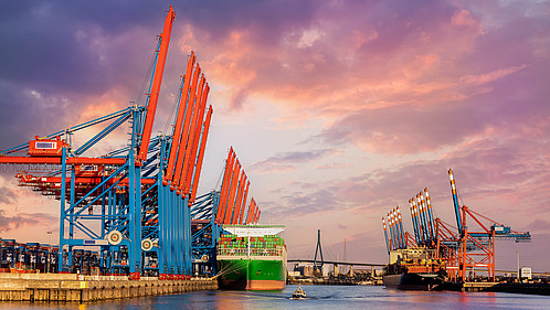 Scenic front giant cargo container ship loading Hamburg city port harbour seaport cranes warm dramatic sky evening sunset light. Global commercial trade freight charter shipping logistics background.