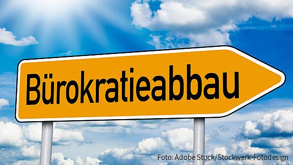 Gelbes, pfeilförmiges Straßenschild mit der Aufschrift „Bürokratieabbau“ vor blauem Himmel mit weißen Wolken. Das Schild zeigt nach rechts und symbolisiert den Weg zu weniger Bürokratie.