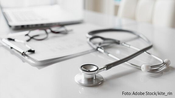 Ein Stethoskop liegt auf einem weißen Schreibtisch vor einem Klemmbrett mit Unterlagen. Daneben befinden sich eine Brille und ein geöffneter Laptop. Die Aufnahme ist hell gestaltet, der Fokus liegt auf dem Stethoskop, während der Hintergrund unscharf ist.