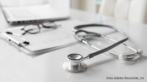 Ein Stethoskop liegt auf einem weißen Schreibtisch vor einem Klemmbrett mit Unterlagen. Daneben befinden sich eine Brille und ein geöffneter Laptop. Die Aufnahme ist hell gestaltet, der Fokus liegt auf dem Stethoskop, während der Hintergrund unscharf ist.