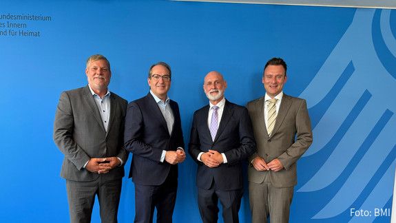 Vier Männer in Anzügen stehen vor einer blauen Wand und blicken in die Kamera