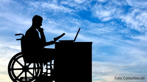 Inklusion und Teilhabe Die Silhouette eines Mannes im Rollstuhl, der an einem Schreibtisch sitzt und arbeitet.