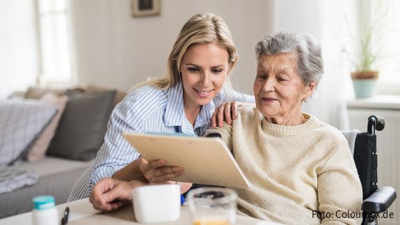 Tag der pflegenden Angehörigen Eine junge Frau sitzt mit einer älteren Frau im Rollstuhl am Esstisch und hilft ihr am Tablet.