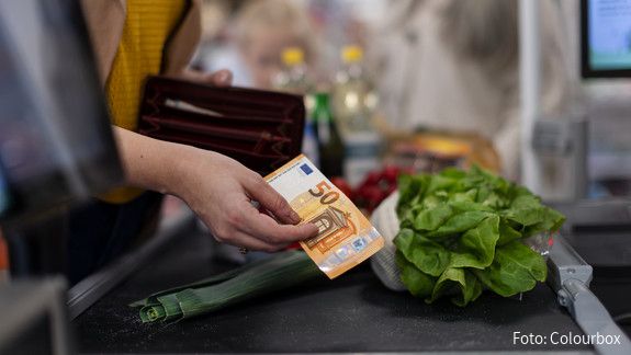 Inflation An einer Supermarktkasse reicht eine Hand einen Geldschein Richtung Kasse, um zu bezahlen. Auf dem Kassenband liegen Lebensmittel.