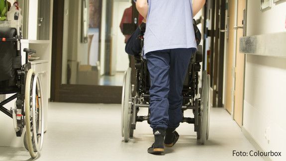 Pflege Eine Pflegekraft, von hinten zu sehen, schiebt einen älterne Menschen im Rollstuhl über einen Flur .