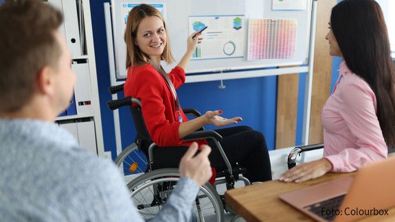Inklusiver Arbeitsmarkt Ein im Rollstuhl sitzende Frau hält vor Kolleginnen und Kollegen einen Vortrag und zeigt auf ein Flipchart, auf dem diverse Grafiken angepinnt sind.