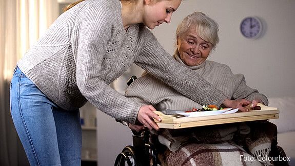 Das Foto zeigt eine Frau, die sich um eine Angehörige kümmert. Sie serviert Essen auf ein Tablett am Rollstuhl.