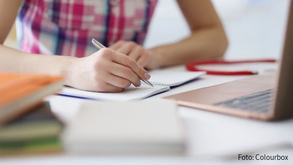 Das Foto zeigt die Hand einer Person, die mit einem Stift auf einem Block schreibt. Rechts im Bild steht ein aufgeklapper Laptop.