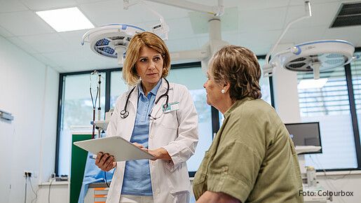 Eine Ärztin steht in einem Krankenzimmer und hält mit ernster Miene ein Klemmbrett
