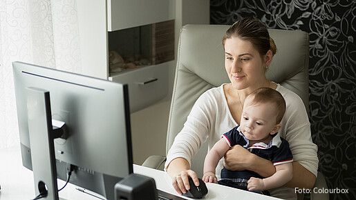 Equal Care Day 2026 Zu sehen ist eine junge Mutter, die am Schreibtisch Homeoffice macht und ihren kleinen Sohn auf dem Arm hält.