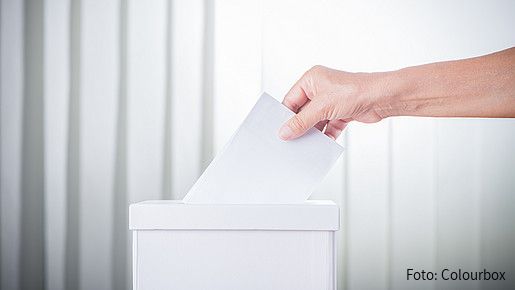 Foto zum Thema "einmal Wahlhelfer, immer Wahlhelfer": Zu sehen ist eine Wahlurne und die Hand einer Person, die gerade ihre Stimme einwirft.