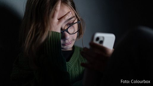 Foto zum Thema "Kinder- und Jugendtelefon": Zu sehen ist ein Mädchen, das in der Dunkelheit sitzt und verzweifelt aufs Smartphone schaut.