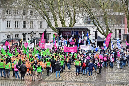 Eine Menschengruppe protestiert mit bunten Fahnen auf einem öffentlichen Platz.
