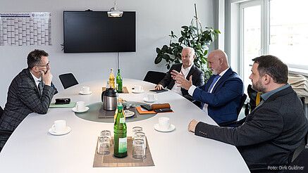 Vier Männer in Anzügen sitzen an einem Konferenztisch und diskutieren