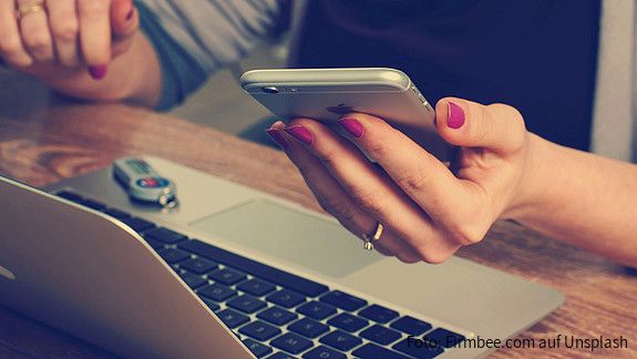 Eine Person sitzt vor einem Laptop und hat ein Handy in der Hand