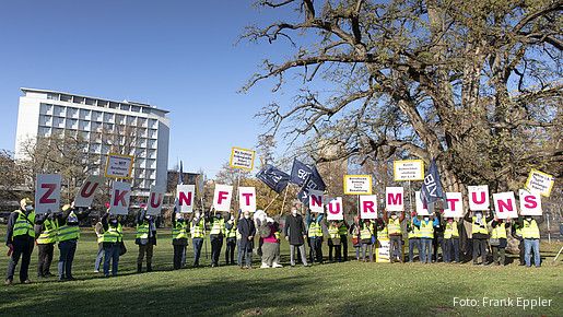 Eine Gruppe von Menschen steht auf einer Grünfläche und protestiert. Sie halten Schilder hoch die zusammen gesetzt "Zukunft nur mit uns" darstellen 