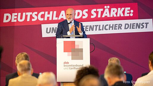 Ein Mann in Anzug steht in einem Konferenzraum hinter einem Rednerpult und spricht in das Mikrofon