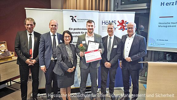 Sechs Personen stehen bei einer Preisverleihung nebeneinander vor den Bannern der Rektorenkonferenz der Hochschulen für den öffentlichen Dienst und der Hessischen Hochschule für öffentliches Management und Sicherheit. Die Person in der Mitte hält eine Urkunde zum 1. Preis sowie eine Rose in den Händen.