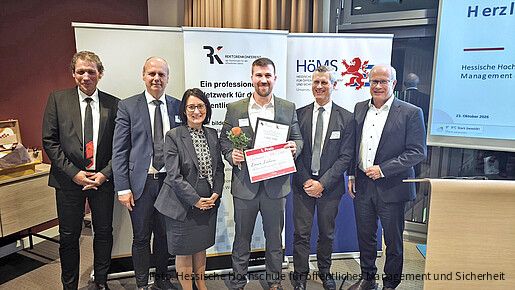 Sechs Personen stehen bei einer Preisverleihung nebeneinander vor den Bannern der Rektorenkonferenz der Hochschulen für den öffentlichen Dienst und der Hessischen Hochschule für öffentliches Management und Sicherheit. Die Person in der Mitte hält eine Urkunde zum 1. Preis sowie eine Rose in den Händen.