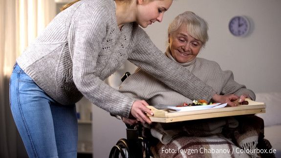 Tag der pflegenden Angehörigen Eine junge Frau reicht einer älteren Frau im Rollstuhl ein Tablett mit einem Mittagessen.