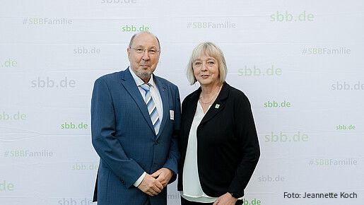 Eine Frau und ein Mann in formeller Kleidung stehen vor einer grauen Leinwand und blicken in die Kamera