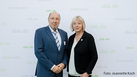 Eine Frau und ein Mann in formeller Kleidung stehen vor einer grauen Leinwand und blicken in die Kamera