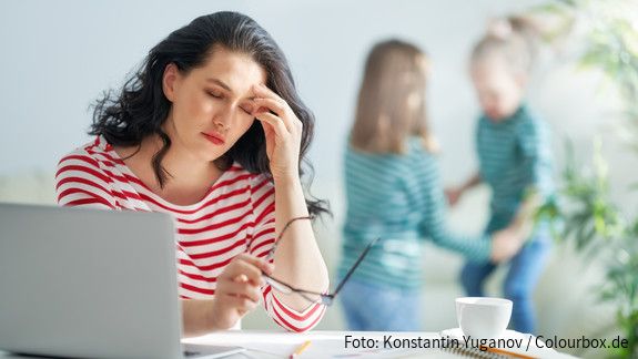 Eine dunkelhaarige Frau arbeitet am Laptop und reibt sich gestresst die Augen, im Hintergrund spielen zwei kleine Mädchen