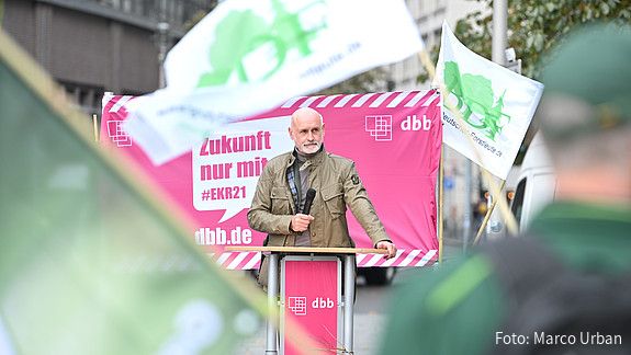 Ein Mann steht im Mittelpunkt des Bildes und spricht in ein Mikrofon