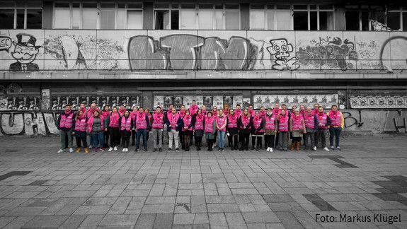 Eine Gruppe junger Beschäftigter des öffentlichen Dienstes in Warnwesten steht vor einem abrissreifen Plattenbau