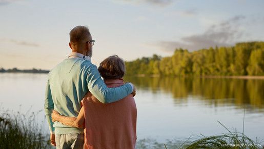 Das Foto zeigt ein Paar (Senioren), die am Wasser stehen und zuversichtlich in Richtung Horizont blicken.