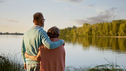 Das Foto zeigt ein Paar (Senioren), die am Wasser stehen und zuversichtlich in Richtung Horizont blicken.