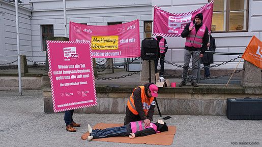 Eine Gruppe von Menschen ist am protestieren. Im Mittelpunkt übt jemand eine Herzdruckmassage an einer Puppe aus. Daneben ist ein Plakat zu sehen, auf dem steht: Wenn uns die TdL nicht langsam beatmet geht uns die Luft aus!