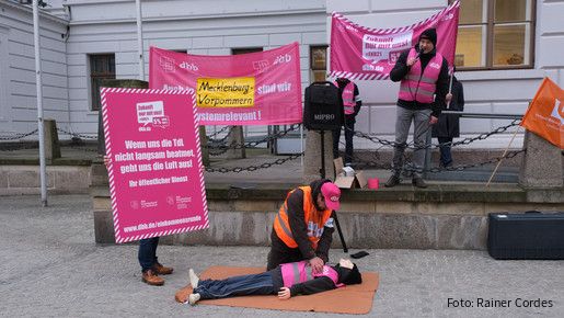 Eine Gruppe von Menschen ist am protestieren. Im Mittelpunkt übt jemand eine Herzdruckmassage an einer Puppe aus. Daneben ist ein Plakat zu sehen, auf dem steht: Wenn uns die TdL nicht langsam beatmet geht uns die Luft aus!