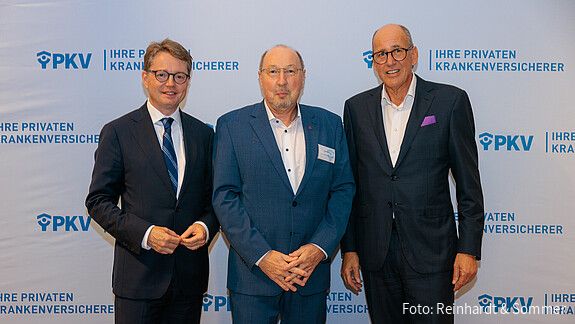 Drei Männer in Anzügen stehen vor einer weißen Leinwand mit PKV-Logos und blicken in die Kamera