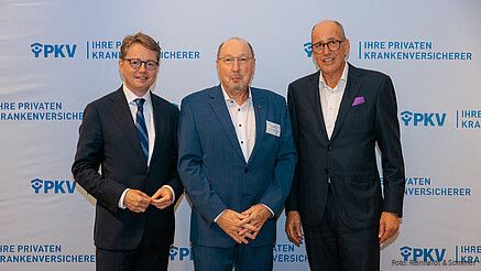 Drei Männer in Anzügen stehen vor einer weißen Leinwand mit PKV-Logos und blicken in die Kamera