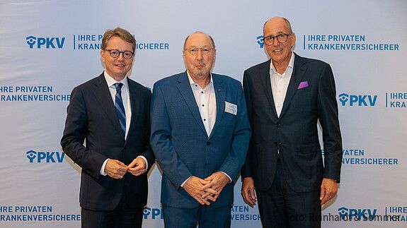 Drei Männer in Anzügen stehen vor einer weißen Leinwand mit PKV-Logos und blicken in die Kamera