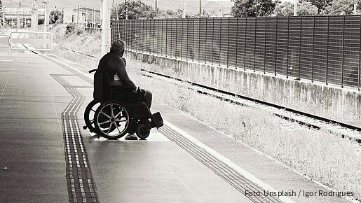 Schwarz-weiß-Foto eines Mannes, der an einem Bahnsteig in einem Rollstuhl sitzt und auf den Zug wartet.