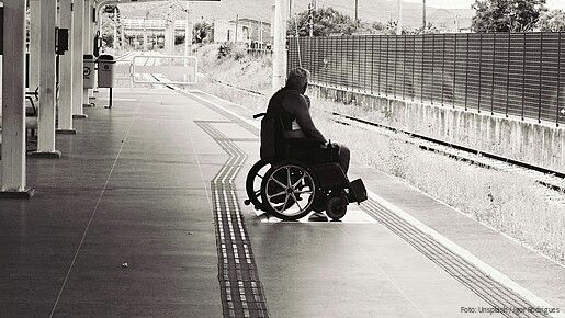 Schwarz-weiß-Foto eines Mannes, der an einem Bahnsteig in einem Rollstuhl sitzt und auf den Zug wartet.