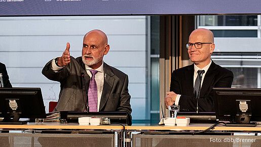 Zwei Männer in Anzügen sitzen an einem Tisch uns sprechen in ein Mikrofon