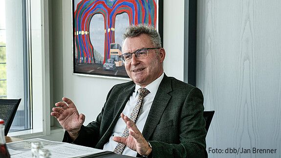 Ein Mann sitzt an einem Schreibtisch und gestikuliert in die Kamera