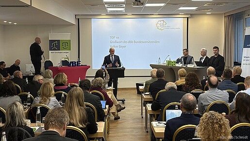 Blick von hinten in einen Konferenzsaal. Im Vordergrund sieht man das Publikum, im Hintergrund steht ein Mann an einem Rednerpult und spricht in ein Mikrofon.