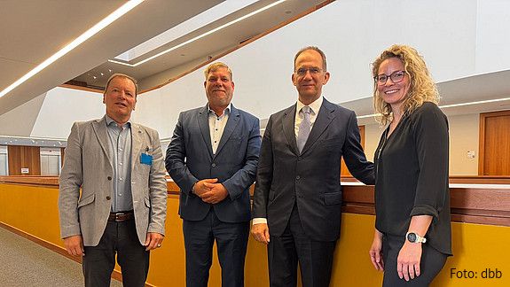 Drei Männer und eine Frau stehen in einer Reihe in einem Büroflur und blicken in Richtung Kamera