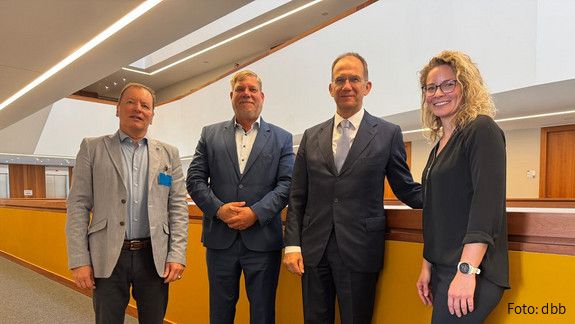 Drei Männer und eine Frau stehen in einer Reihe in einem Büroflur und blicken in Richtung Kamera