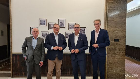 Vier Männer in Anzügen stehen vor einer Wand mit Bildern und blicken in die Kamera