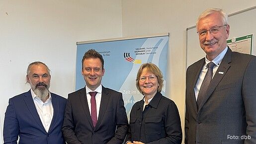 Drei Männer in Anzügen und eine Frau mit Bluse stehen vor einem hellblauen Aufsteller und blicken in die Kamera.