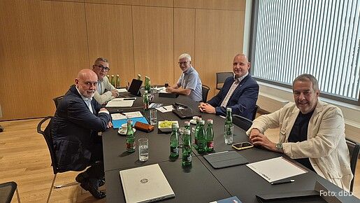 Fünf Personen sitzen an einem Konferenztisch in einem Besprechungsraum. Auf dem Tisch stehen Wasserflaschen, Gläser, Unterlagen und Laptops.