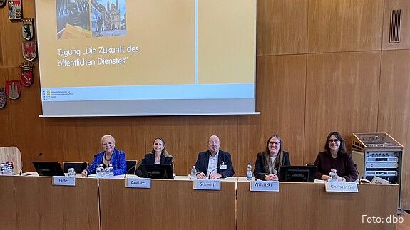 Vier Frauen und ein Mann sitzen an einem langen Rednerpult und blicken in die Kamera