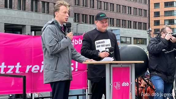 Zwei junge Männer stehen auf einem Podest auf einer Demonstration, der linke der beiden, Florian Schütz, spricht in ein Mikrofon. 